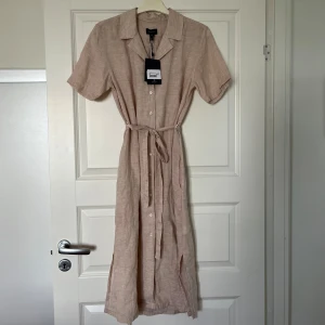 Beige linneklänning från Gant - Säljer en beige skjortklänning från Gant i linne. Klänningen har korta ärmar, knappar hela vägen framtill och ett matchande knytskärp i midjan (som kan tas bort). Det är en midiklänning i storlek 36. 
