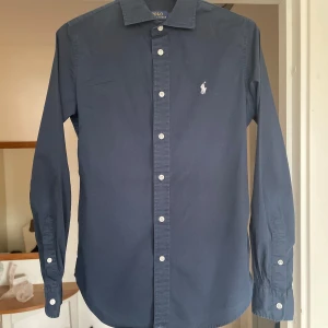 Mörkblå skjorta från Polo Ralph Lauren - Snygg mörkblå skjorta från Polo Ralph Lauren med klassisk krage och vita knappar. Skjortan har lång ärm och den ikoniska broderade loggan på bröstet. Perfekt till jeans eller chinos för en stilren look. Sjukt skön skjorta inför sommaren! 