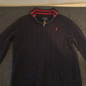 Mörkblå kabelstickad tröja med dragkedja från Polo Ralph Lauren - Säljer en mörkblå stickad tröja från Polo Ralph Lauren med halv dragkedja och röd logga på bröstet.  Tröjan är köpt på NK men säljer nu för den är för liten. Nypris 1500