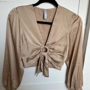 Beige omlottopp med knyt från A M I S U - Säljer en beige omlottopp från A M I S U i satinliknande material. Toppen har lång ärm, djup v-ringning och knyt framtill med dekorativ ring. Perfekt för dig som gillar trendiga detaljer och en elegant look.