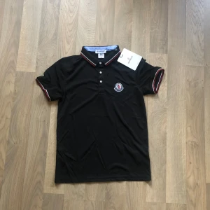 Svart piké från Moncler - Svart pikétröja från Moncler med klassisk krage och knäppning framtill. Tröjan har Moncler-logga på bröstet och snygga rödvita detaljer på krage och ärmslut. Perfekt för en stilren och sportig look. Storleken är M men passar även S lika bra. Säljer den pga jag vill ändra stilen, har bara använt den ett par gånger. Pris kan diskuteras vid snabb affär. Byten funkar också!