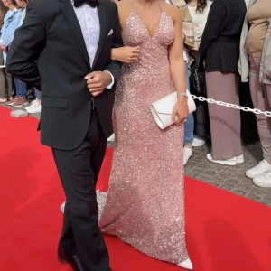 Glittrig rosa balklänning med öppen rygg - Säljer en lång, glittrig rosa balklänning med tunna axelband och djup v-ringning. Klänningen har öppen rygg och är täckt av paljetter som ger ett riktigt lyxigt intryck. Perfekt för bal eller fest där du vill sticka ut och glänsa. den är ifrån DRESSROOM LINNE. ORGINALPTIS 5000kr!! En liten del av paljetterna har gått sönder där fram men finns en påse med paljetter ( de syns inte när man kollar på klänningen) därav pris  