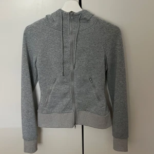 Grå hoodie med dragkedja och huva - Snygg grå hoodie med hel dragkedja, huva med snörning och två fickor med dragkedja. Tröjan har långa ärmar med tumhål och breda muddar. Perfekt för chill eller träning, i mjukt bomullsmaterial.