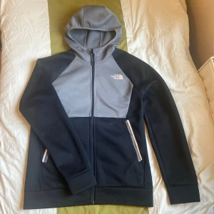 The North Face Svart och grå kofta - Snygg kofta från The North Face i svart och grått. Jackan har dragkedja framtill, två fickor med dragkedja och en huva. Perfekt för chill dagar eller när du vill ha en sportig look. Tillverkad i mjukt material för extra komfort. Pris kan diskuteras 