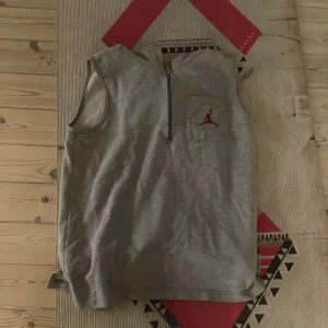 Grå Jordan väst med huva och half zip - Säljer en grå ärmlös väst från Jordan med huva och half zip framtill. Västen har en bröstficka med röd Jordan logga och är gjord i mjukt bomullsmaterial. Perfekt för en sportig och avslappnad stil.