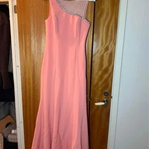 Lång rosa balklänning med strassdetaljer - Säljer en elegant långklänning i rosa med oneshoulder-design och transparent mesh upptill. Klänningen har glittriga strassdetaljer längs kanten och är perfekt för bal eller fest. Materialet är slätt och följsamt.