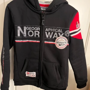 Svarte hoodie från Geographical Norway - Svart hoodie med dragkedja från Geographical Norway. Tröjan har tryck och broderade detaljer i vitt, rött och grått samt flaggor på ärmen. Två fickor framtill och ribbade muddar. Perfekt för en sportig stil.  