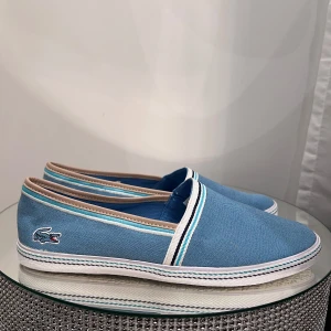 Sneakers  - Snygga ljusblå slip-on sneakers från Lacoste med vit sula och diskreta ränder längs kanten. Skorna har det klassiska Lacoste-krokodilen broderad på sidan och insida med randigt tyg. Perfekta för en avslappnad stil.