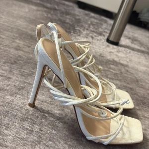 Vita klackar med snörning - Säljer ett par vita sandaler med hög klack och fyrkantig tå. Skorna har snörning som går att vira runt benet och ett diskret krokodilpräglat mönster. Perfekta för dig som vill sticka ut med stilrena detaljer. ALDRIG ANVÄNDA