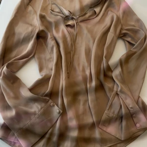 Guldig blus med knytning från H&M - Säljer en guldig blus från H&M med långa ärmar och knytning vid halsen. Blusen har en lätt glansig yta och knappar vid ärmsluten. Perfekt för dig som gillar en stilren och trendig look.