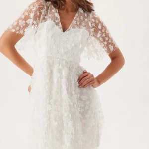 Vit blommig kortklänning med spetsdetaljer - Superfin vit kortklänning med transparenta ärmar och överdel täckt av blommiga spetsdetaljer. Klänningen har v-ringning och en lätt utsvängd kjol. Aldrig använd med prislappen kvar. Perfekt för sommarens speciella tillfällen.