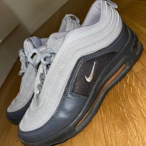 Skor - Säljer ett par Nike Air Max 97 modell (ej äkta märke) i grått och svart med vita detaljer. Skorna har den klassiska vågiga designen, synlig luftbubbla i sulan och snörning framtill. Perfekta för dig som gillar streetwear och sportig stil.