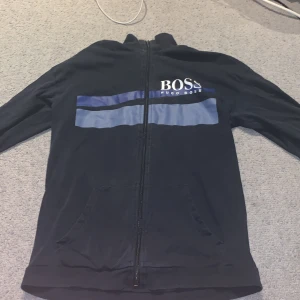 Mörkblå zip-hoodie från Hugo Boss - Säljer en mörkblå hoodie med dragkedja från Hugo Boss. Tröjan har två blåa ränder och stor vit logga på bröstet. Praktiska fickor framtill och detaljerad dragkedja med Boss-gravyr. Perfekt för en stilren och avslappnad look.