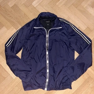 Mörkblå vindjacka från Smog - Säljer en mörkblå vindjacka från Smog med vita ränder längs ärmarna och dragkedja framtill. Jackan har en sportig look med elastiska muddar och är perfekt för en casual stil. Passar bra för vår och höst.