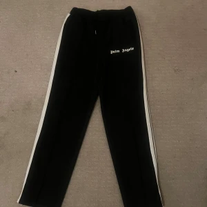 Svarta byxor från Palm Angels - Snygga svarta byxor från Palm Angels med vita sidoremsor och logga på framsidan. De har en elastisk midja med snörning för extra komfort. Perfekta för en avslappnad stil.