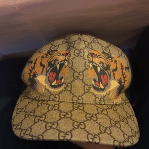 Gucci keps med tigertryck - Snygg keps från Gucci med ett djärvt tigertryck på framsidan och det ikoniska GG-mönstret i bakgrunden. Kepsen har en justerbar rem baktill med de klassiska Gucci-färgerna. 