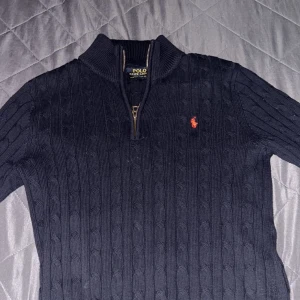 Svart stickad tröja från Ralph Lauren - Säljer en stilren svart stickad tröja från Ralph Lauren med dragkedja och broderad logga på bröstet. Tröjan har en slim fit passform och långa ärmar, perfekt för kyligare dagar.