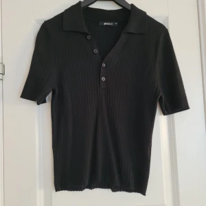 Svart ribbad pikétröja från Gina tricot - Snygg svart ribbad pikétröja från Gina med korta ärmar och knappar framtill. Perfekt för en stilren look. Passar bra till både jeans och kjol.