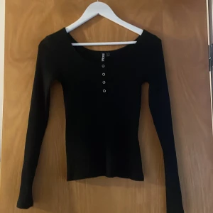 Svart ribbad tröja från Pieces - Snygg svart ribbad tröja från Pieces med lång ärm och knapprad framtill. Tröjan har ett litet hål längst ner på armen men som knappt syns tycker jag. 