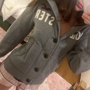 Grå hoodie från Hollister - Säljer en grå hoodie från Hollister med knappar framtill och justerbar huva. Tröjan har en broderad logga och fickor på sidorna. Perfekt för en avslappnad stil.