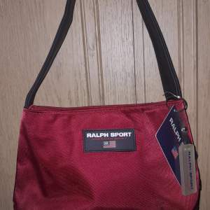 Snygg röd axelväska från Ralph Lauren Sport med svart axelrem. Väskan har en stilren design med en logotyp på framsidan. Perfekt för att bära dina dagliga nödvändigheter med stil.