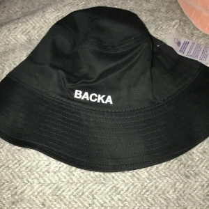 Svart bucket hat från Atlantis - Snygg svart bucket hat från Atlantis med texten 'BACKA' tryckt på framsidan. Tillverkad i bomull för en bekväm passform. Perfekt för en avslappnad stil. Denna är ny med prislapp och en ny från butik kostar 179kr med 10% rea