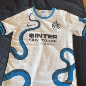 Vit Inter Milan fotbollströja - Säljer en vit Inter Milan fotbollströja från Nike med blått ormmönster. Tröjan har korta ärmar och sponsorloggor från $INTER Fan Token och Lenovo. Perfekt för fans av laget!