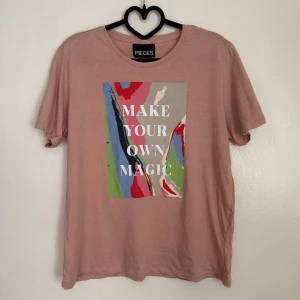 Rosa t-shirt från Pieces - Säljer en rosa t-shirt från Pieces med trycket 'Make Your Own Magic' i vitt. T-shirten har ett färgglatt abstrakt mönster i blått, rött och grönt på framsidan. Perfekt för en avslappnad stil.