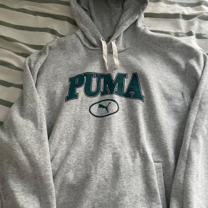 Grå hoodie från Puma - Säljer en grå hoodie från Puma med stor logga i grönt och svart på bröstet. Perfekt för en avslappnad stil. Hoodien är i storlek L men passar nog även de flesta med storlek M.