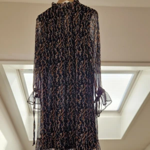 Mönstrad långklänning med volanger - Säljer en elegant långklänning med ett unikt mönster i mörka och ljusa toner. Klänningen har långa ärmar med volangdetaljer och knytband vid ärmsluten. Perfekt för en stilfull look.