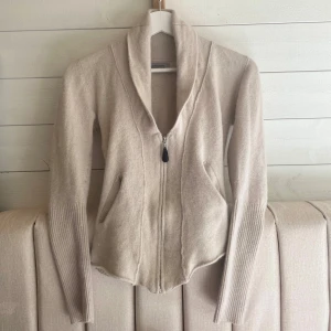 Beige kofta från HunkyDory Essentials - Säljer en stilren beige kofta från HnkyDory Essentials. Den har en dragkedja framtill och långa ärmar med ribbade detaljer vid ärmsluten. Perfekt för en avslappnad look! 70% Lammull, 20% Angora så den är jättemjuk!