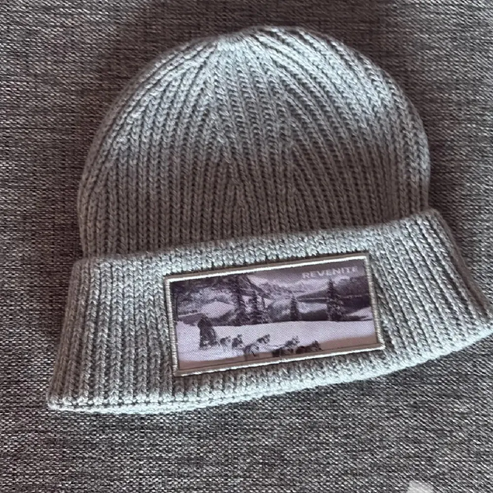 Snygg grå stickad mössa från Revenite med ett coolt patchmotiv av ett vinterlandskap och hundspann. Perfekt för kyliga dagar.. Asusteet.