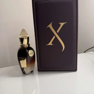 Lyxig parfym från Xerjoff - Exklusiv parfym från Xerjoff i en elegant flaska med guldiga detaljer och en distinkt X-logga. Flaskan har en mörk toning och kommer i en stilren lila förpackning med guldtext. Perfekt för den som söker en sofistikerad doftupplevelse.