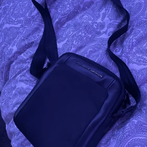 Svart axelväska från Porsche Design - Snygg och stilren svart axelväska från Porsche Design. Väskan har en justerbar axelrem och en praktisk dragkedja för säker förvaring. Perfekt för att bära med sig det nödvändigaste med stil.