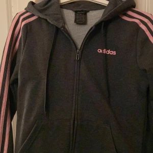Svart/grå hoodie från Adidas med rosa detaljer - Säljer en snygg svart hoodie från Adidas med rosa ränder på ärmarna och rosa logga på bröstet. Den har en dragkedja framtill och en praktisk huva med snörning. Perfekt för en sportig look eller en avslappnad dag.