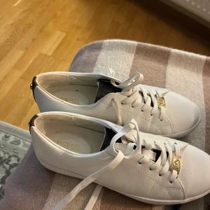 Vita sneakers från Michael Kors - Snygga vita sneakers från Michael Kors med gulddetaljer och snörning. Skorna har en stilren design med märkets logotyp på sidan och baksidan. Perfekta för en trendig look.