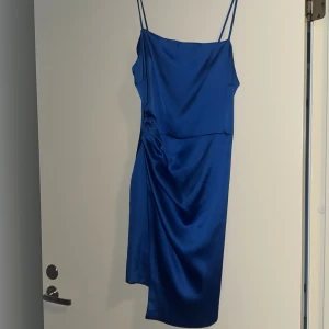 Blå sidenklänning med smala axelband - Snygg blå klänning i siden med smala axelband och en elegant drapering på sidan. Perfekt för en kväll ute eller en speciell tillställning. Klänningen har en figurnära passform som framhäver silhuetten.