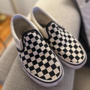Svartvita rutiga slip-on sneakers från Vans - Säljer ett par klassiska svartvita slip-on sneakers från Vans med ikoniskt rutigt mönster. Skorna har en bekväm passform och är perfekta för en avslappnad stil. 