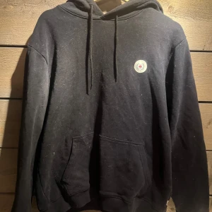 Svart hoodie från Woodbird - Säljer en svart hoodie från Woodbird med en liten logga på bröstet. Den har en klassisk design med dragsko i huvan och en stor magficka. Perfekt för en avslappnad stil.