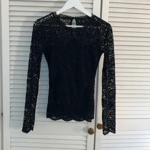Svart spetsblus från Vero Moda - Elegant svart spetsblus från Vero Moda med långärmad design. Blusen har ett vackert spetsmönster som ger en sofistikerad look. Perfekt för att lyfta vilken outfit som helst.