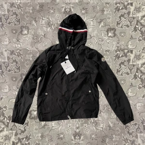 Svart vindjacka från Moncler - Snygg svart vindjacka från Moncler med huva och dragkedja. Jackan har en stilren design med Moncler-logga på ärmen och en randig detalj på huvan. Perfekt för blåsiga dagar!