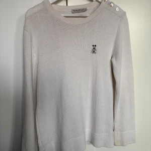 Vit stickad tröja - Säljer en stilren vit stickad tröja med en liten broderad detalj på bröstet. Tröjan har långa ärmar och knappar på axeln för en extra touch. Perfekt för en elegant och avslappnad look.