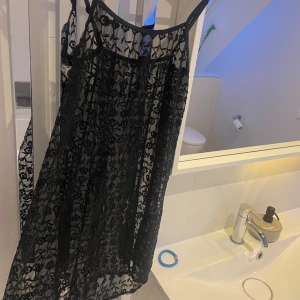 Svart spetsklänning - Säljer en elegant svart spetsklänning med blommigt mönster. Klänningen är ärmlös och har en luftig passform, perfekt för en stilfull look. Passar utmärkt till festliga tillfällen.