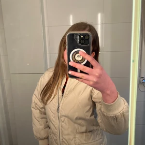 Beige bomberjacka - Superfin vit jacka i storlek 134 från Kappahl! En stilren och klassisk modell som passar perfekt för både vår och höst. Jackan är i bra skick och har mycket kvar att ge. Den är lätt fodrad, vilket gör den lagom varm under kyliga morgnar utan att bli för tung att bära under dagen. Jackan har två praktiska fickor framtill – perfekt för små skatter, vantar eller bara för att värma händerna. Dragkedja framtill och bra passform som gör den bekväm att röra sig i, både till lek och vardag. 