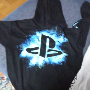 Svart hoodie med PlayStation-tryck - Säljer en svart hoodie med ett coolt PlayStation-tryck i blått och vitt. Perfekt för gamingkvällar eller som en avslappnad vardagströja. Hoodien har en klassisk passform med huva och långa ärmar.