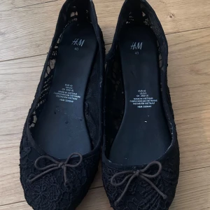 Svarta ballerinaskor i spets från H&M - Snygga svarta ballerinaskor från H&M med spetsdetaljer och en söt rosett framtill. Perfekta för en stilren look.