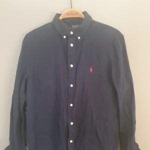 Mörkblå skjorta från Ralph Lauren - Snygg mörkblå skjorta från Ralph Lauren med klassisk krage och knappar framtill. Den har en broderad röd logotyp på bröstet och är långärmad. Perfekt för en stilren look.