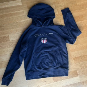 Mörkblå hoodie från GANT - Säljer en jättesnygg mörkblå hoodie från GANT med broderad logga. Tröjan har en klassisk känguruficka och en bekväm huva. Nästan aldrig ancvänd och inga fläckar!!❣️