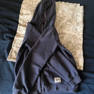 Mörkblå hoodie från Andy - Säljer en mörkblå hoodie från Andy med dragkedja och huva. Den har långa ärmar och ribbade muddar vid ärmslut och nederkant. Perfekt för en avslappnad stil. Finns tecken på användning, som en väldigt vag fläck som inte går bort, syns knappt om man inte kollar väldigt nära och noga. Säljs pga att den blivit för liten för mig, men annars i riktigt bra skick.