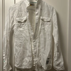 Vit långärmad skjorta med fickor - Säljer en stilren vit långärmad skjorta med två bröstfickor med lock. Skjortan har en klassisk krage och knappar framtill. Perfekt för en avslappnad men ändå uppklädd look.